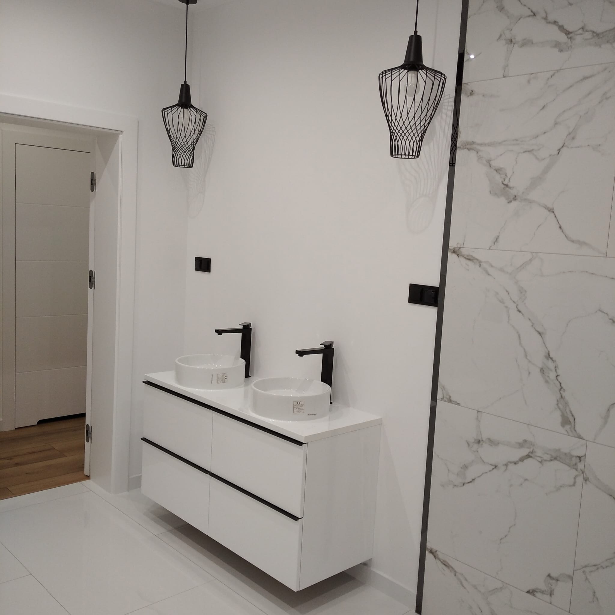 Nowoczesna łazienka z podwójną umywalką na białej szafce, czarne baterie, marmurowe płytki i designerskie lampy wiszące. Minimalistyczny wystrój.