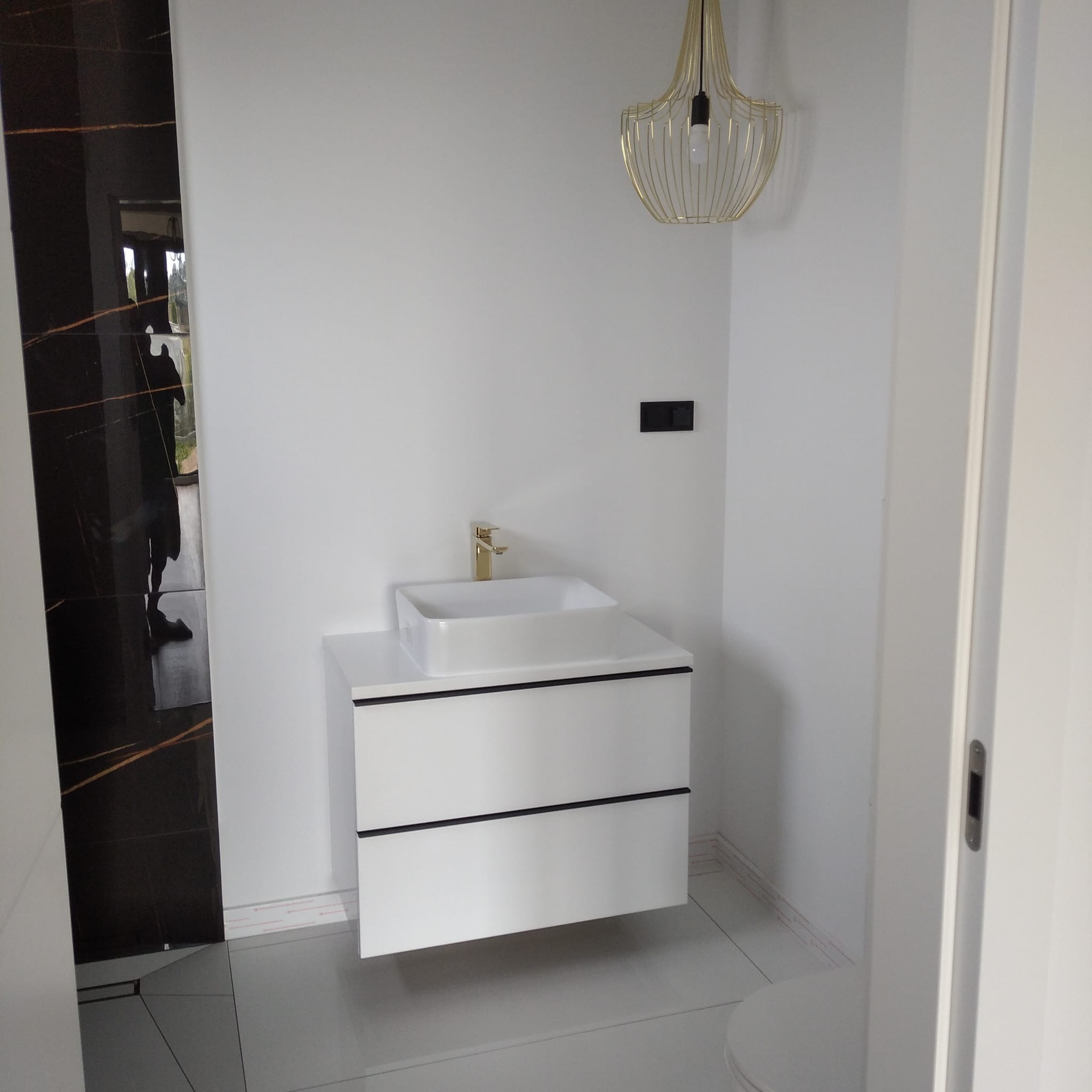 Nowoczesna łazienka z białą szafką pod umywalkę, złotą armaturą i czarną płytką imitującą marmur. Minimalistyczny design, wisząca lampa.