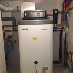 Wojciech woj - Biały, kompaktowy piec Rotex GCU z podłączonymi rurami w kolorze niebieskim i czerwonym, zamontowany w pomieszczeniu z wykończonymi płytkami ścianami.