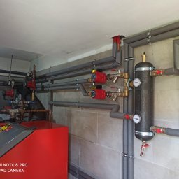 Wojciech woj - Zabezpieczone rury instalacji grzewczej z czerwonymi pompami obiegowymi i czarnym zbiornikiem wyrównawczym, zamontowane na ścianie z płytek w jasnym pomieszczeniu.