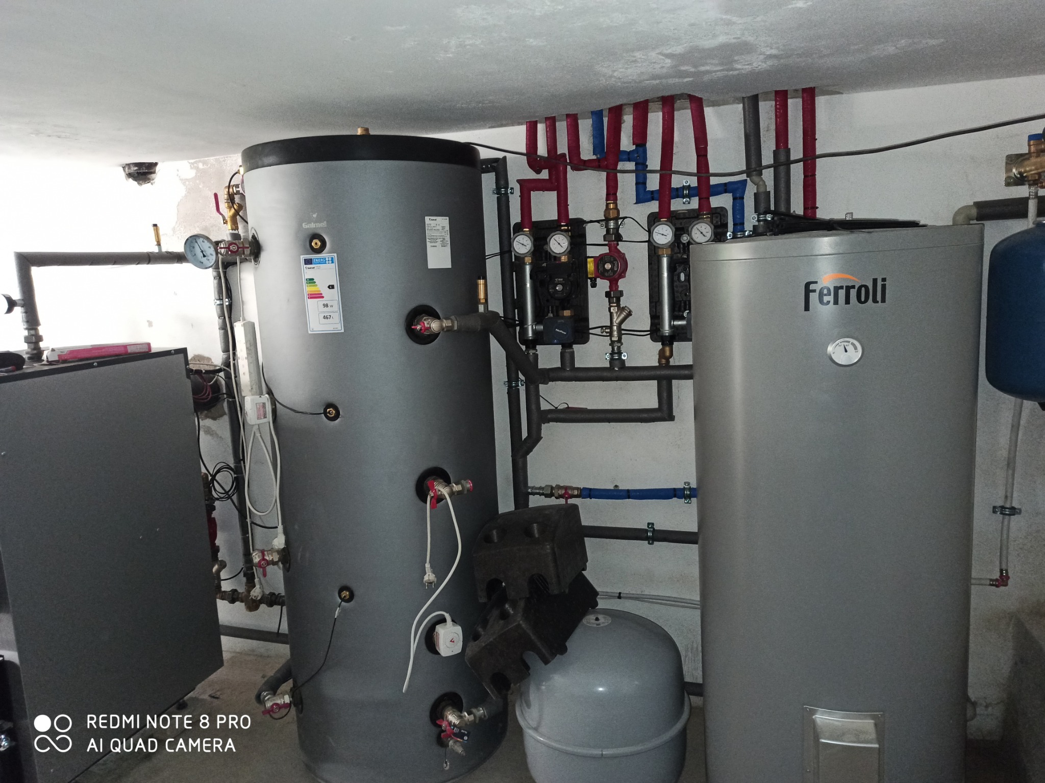 Szary, cylindryczny zbiornik Galmet z etykietą energetyczną, obok zbiornik Ferroli, rury w izolacji, manometry i zawory. Instalacja w kotłowni.