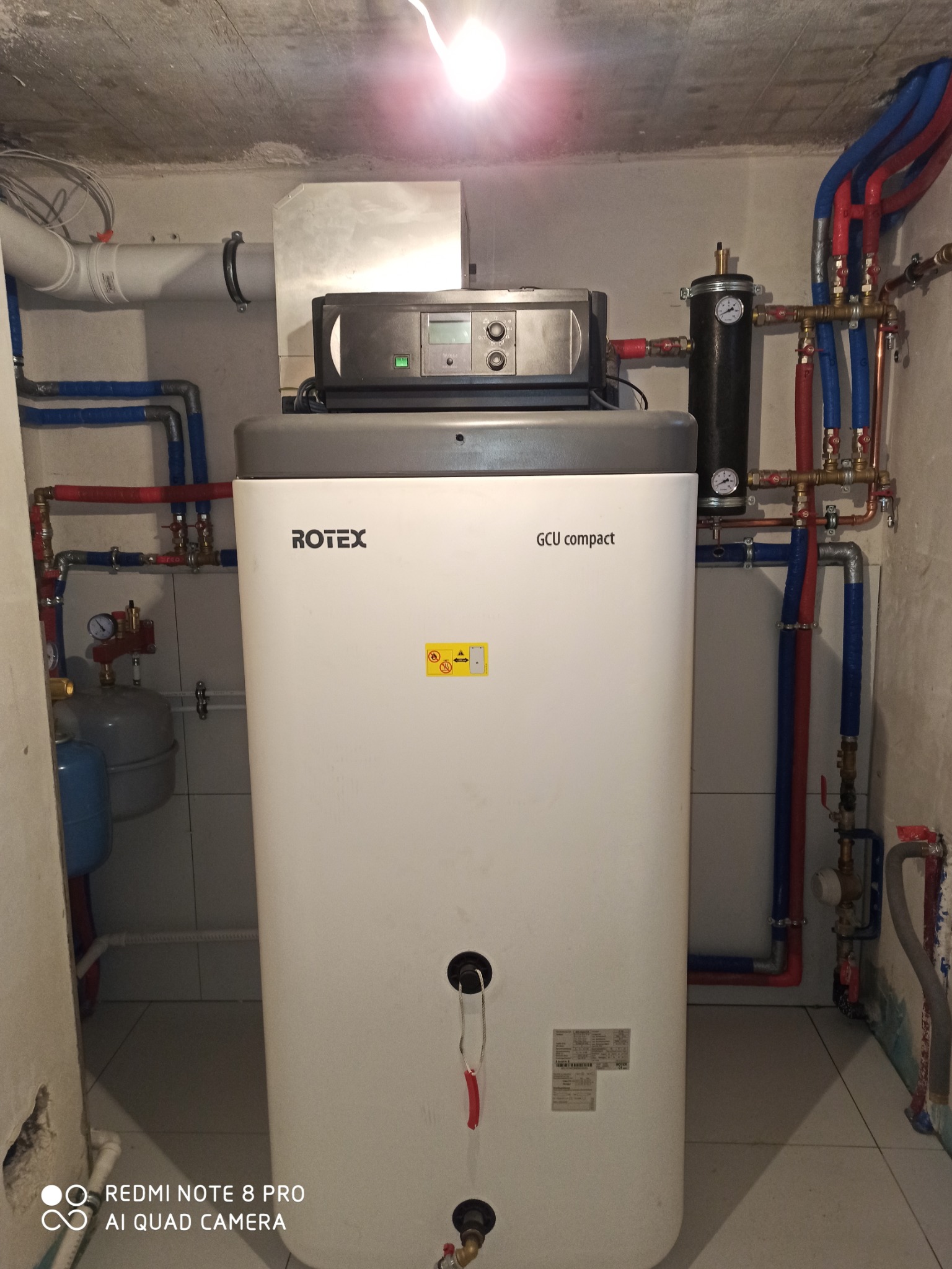 Biały, kompaktowy piec Rotex GCU z podłączonymi rurami w kolorze niebieskim i czerwonym, zamontowany w pomieszczeniu z wykończonymi płytkami ścianami.