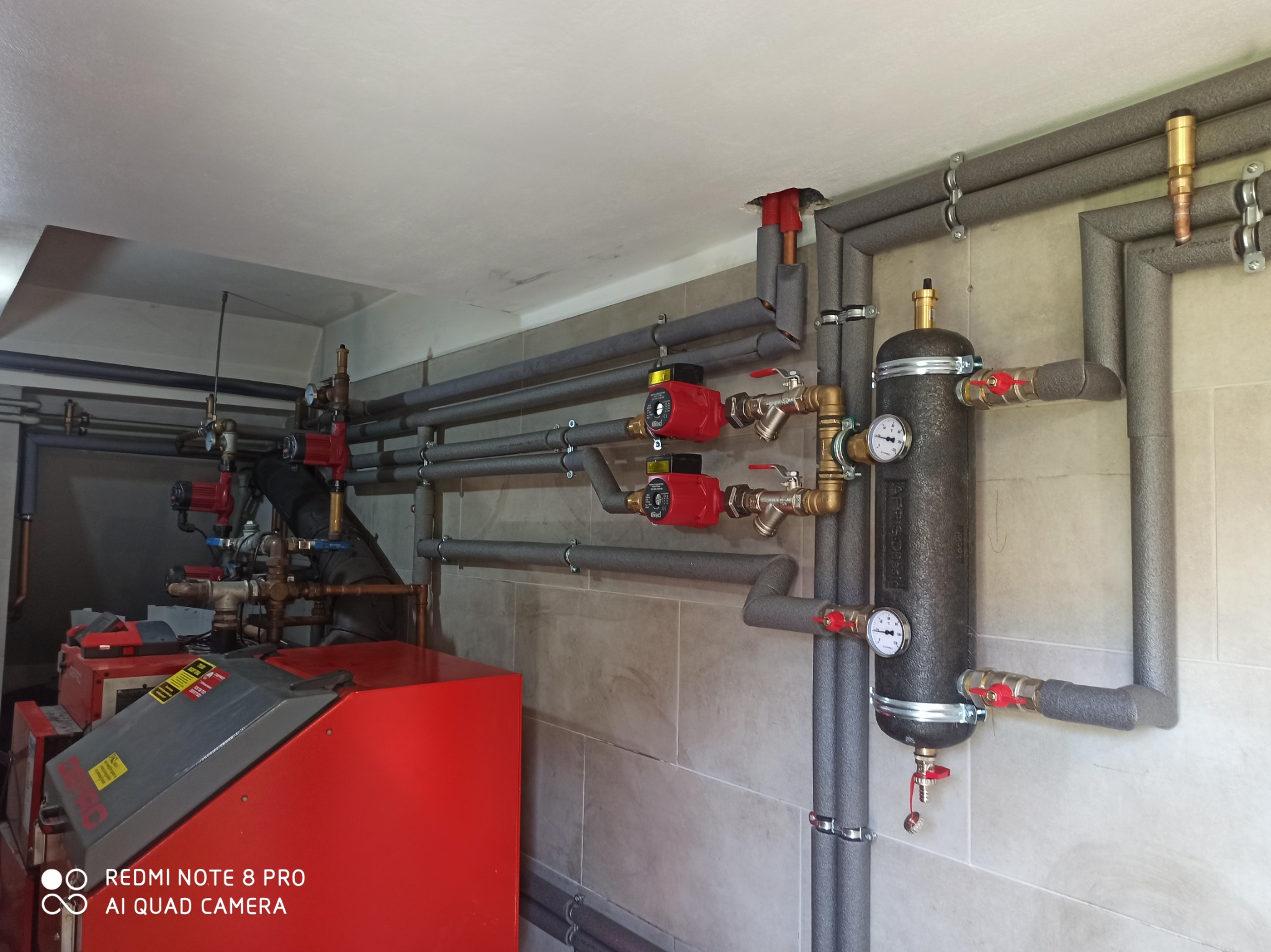 Zabezpieczone rury instalacji grzewczej z czerwonymi pompami obiegowymi i czarnym zbiornikiem wyrównawczym, zamontowane na ścianie z płytek w jasnym pomieszczeniu.