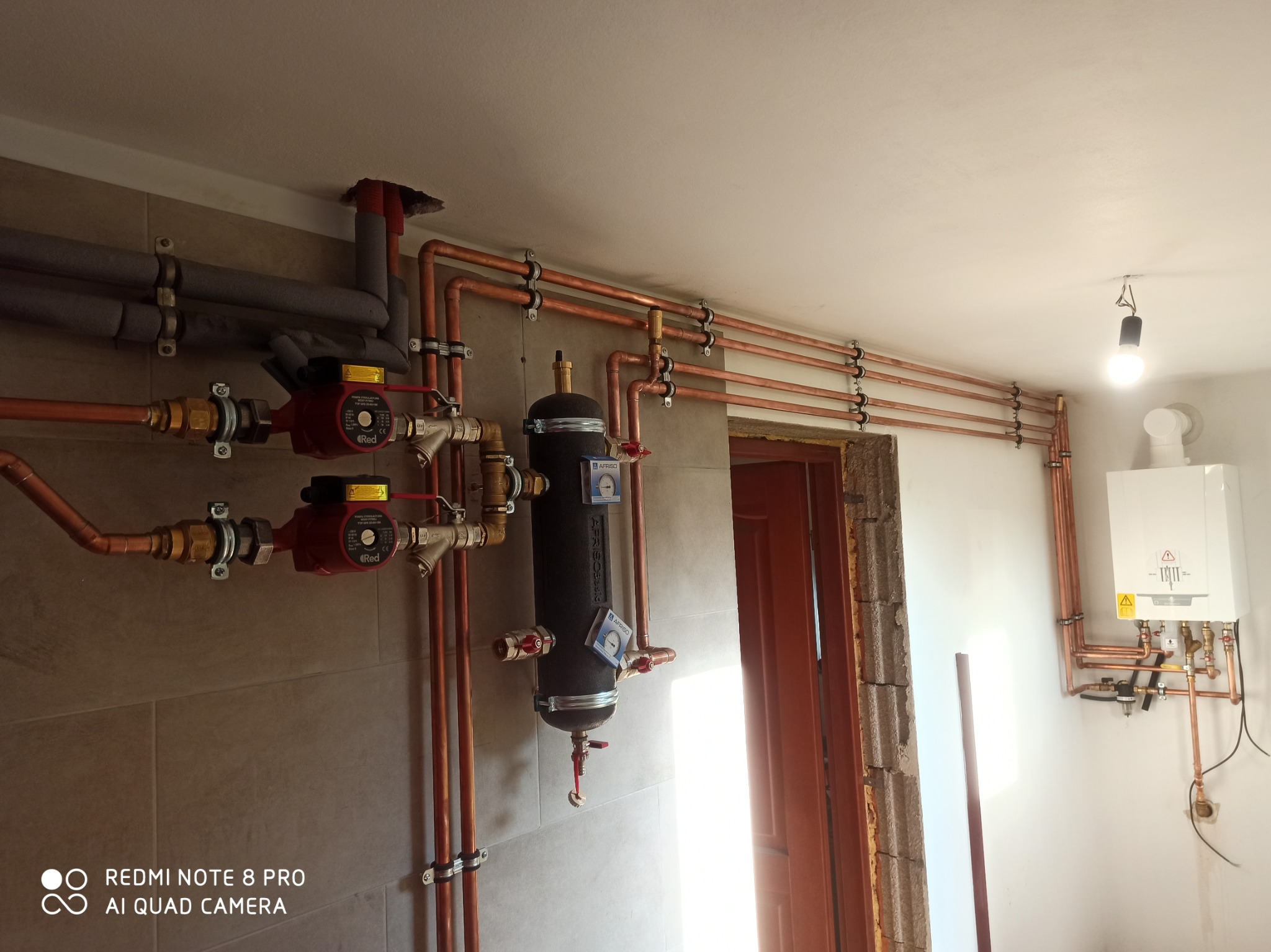 Miedziana instalacja grzewcza z pompami obiegowymi i filtrem, zamontowana na szarej ścianie. Widoczny piec gazowy i rury biegnące pod sufitem.