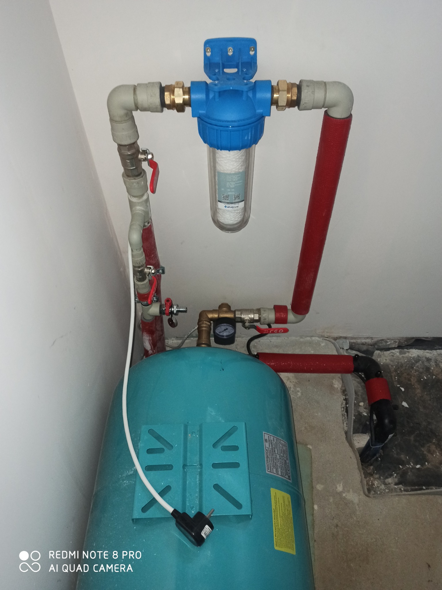 Niebieski filtr wody z przezroczystym wkładem, zamontowany na rurach z zaworami, nad turkusowym zbiornikiem ciśnieniowym w rogu pomieszczenia.