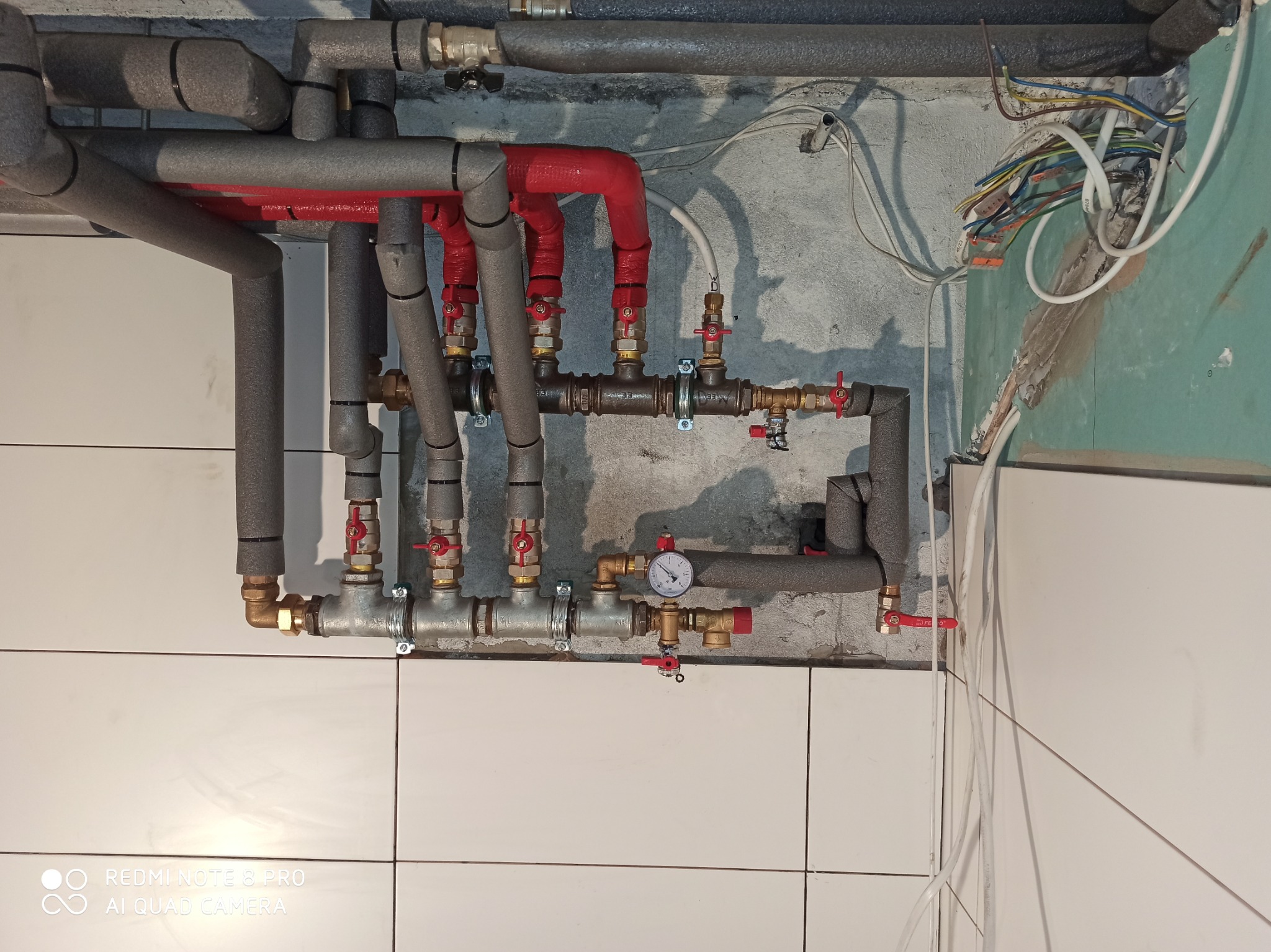 Złożona instalacja hydrauliczna z rurami w izolacji, zaworami i manometrem, zamontowana na ścianie z białymi płytkami i surowym betonem. Widoczne przewody elektryczne.