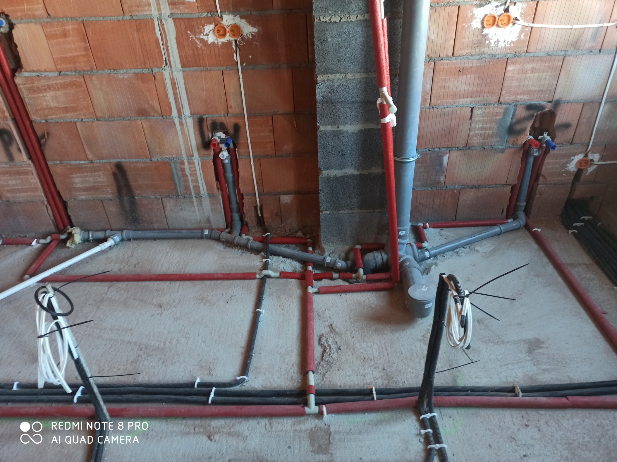 Instalacja wodna i kanalizacyjna w surowym stanie, widoczne rury w kolorze czerwonym, szarym i czarnym, zamocowane do ceglanej ściany i podłoża.