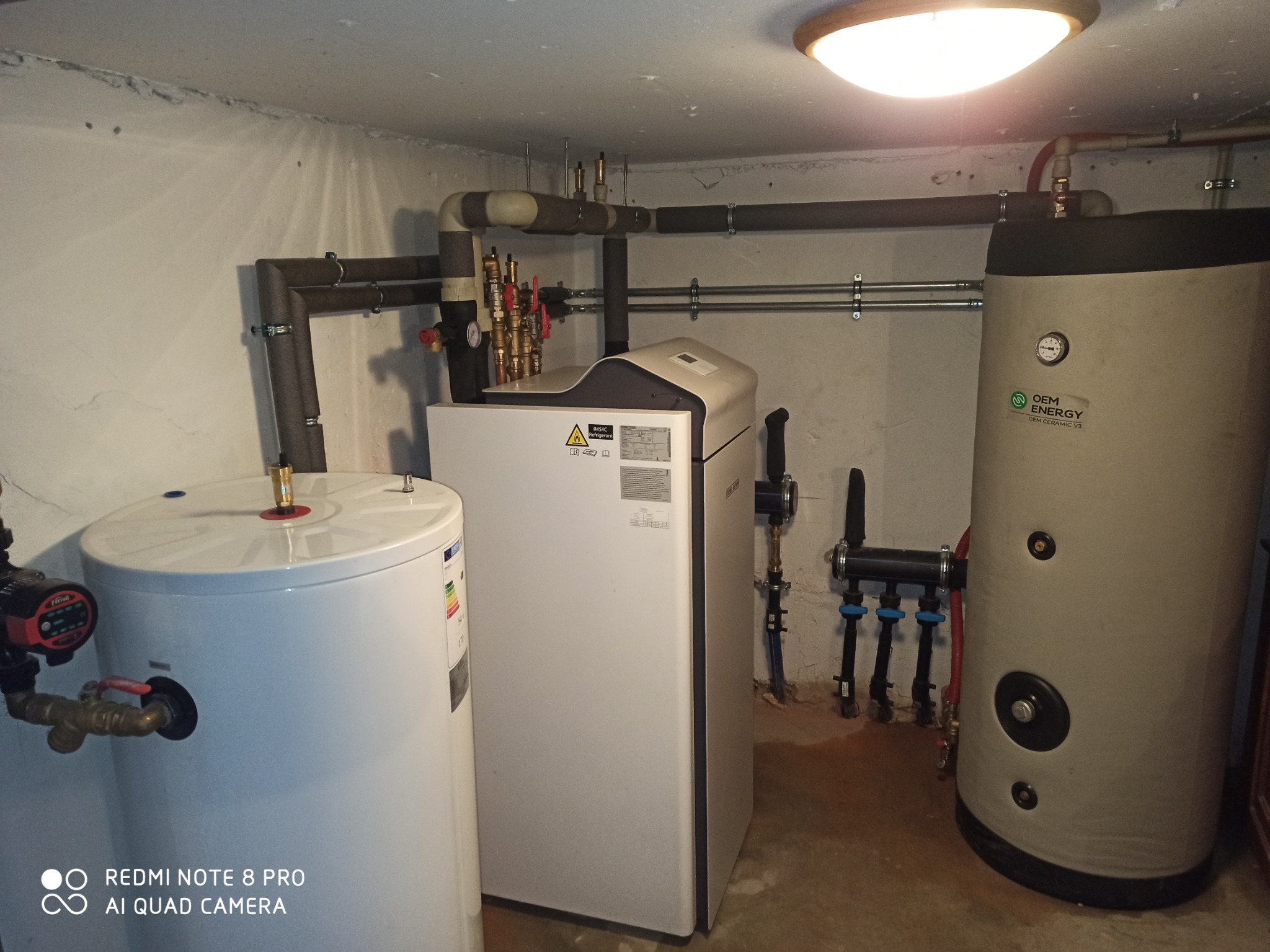 Zestaw urządzeń grzewczych w kotłowni: biały piec, zasobnik ciepłej wody i zbiornik buforowy. Widoczne rury i armatura. Oświetlenie sufitowe.