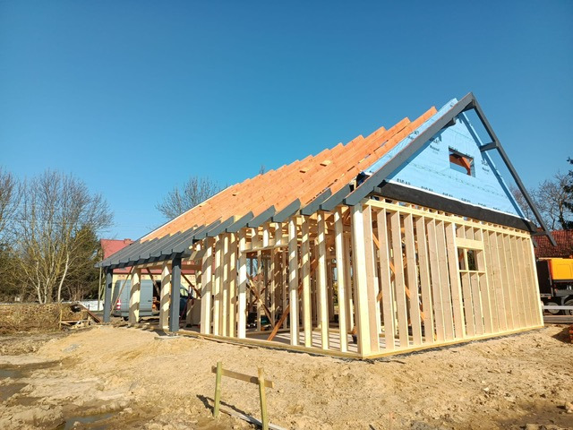 Surowa konstrukcja domu szkieletowego w trakcie budowy, z widoczną drewnianą ramą, częściowo pokrytym dachem i niebieską membraną. Słoneczny dzień, plac budowy.