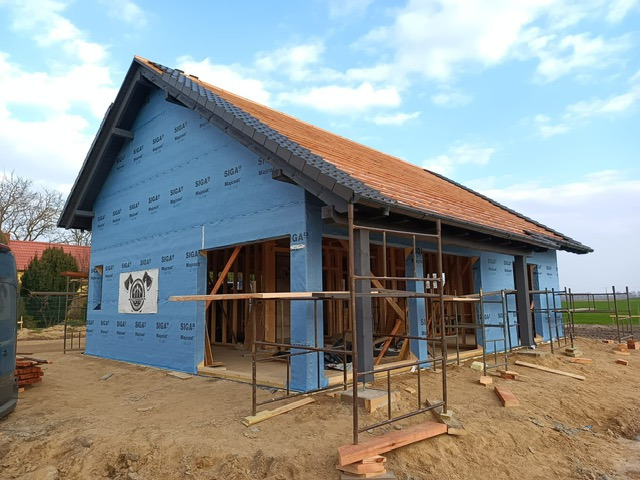 Budowa domu szkieletowego z częściowo położonym dachem z dachówki ceramicznej. Ściany pokryte niebieską membraną. Widoczne rusztowania i drewniana konstrukcja.
