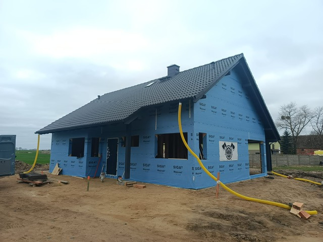Niedokończony dom z niebieską membraną izolacyjną i czarnym dachem. Żółte rury wentylacyjne na zewnątrz. Widoczne okna bez szyb i logo firmy na ścianie.