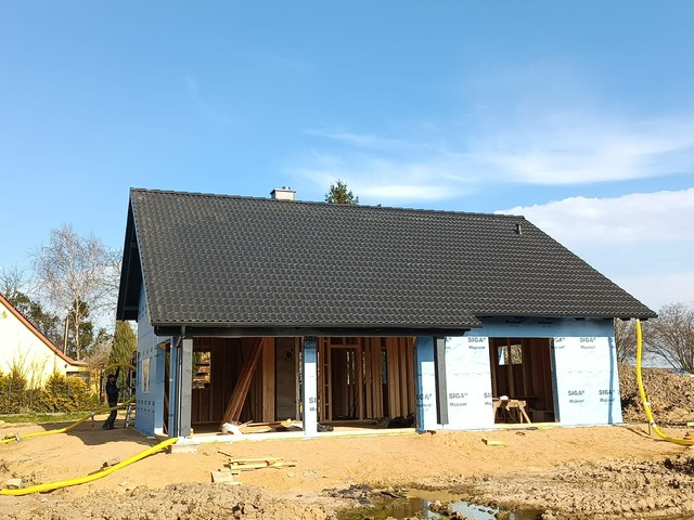 Niedokończony dom szkieletowy z czarnym dachem, widoczne drewniane elementy konstrukcyjne i folia izolacyjna. Żółty wąż na pierwszym planie. Błękitne niebo w tle.