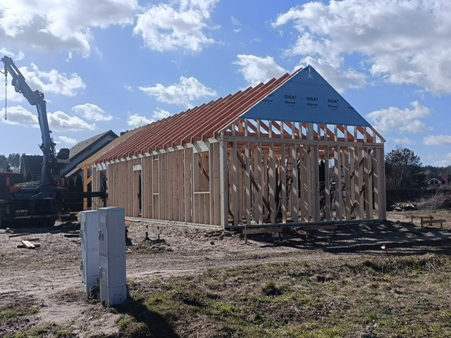 Wznoszenie domu szkieletowego: konstrukcja ścian i dachu z widoczną membraną. Na pierwszym planie skrzynki elektryczne, w tle żuraw budowlany i zabudowa jednorodzinna.