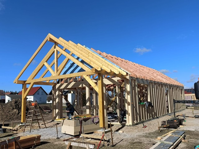 Konstrukcja domu w trakcie budowy: drewniany szkielet z widoczną więźbą dachową i częściowo ułożonymi łatami pod pokrycie. Na pierwszym planie stos desek i drabina.