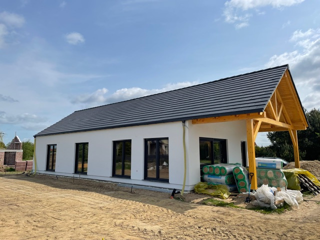 Nowoczesny dom szkieletowy z ciemną dachówką i drewnianą konstrukcją werandy, w trakcie budowy, z widocznymi materiałami izolacyjnymi obok budynku.