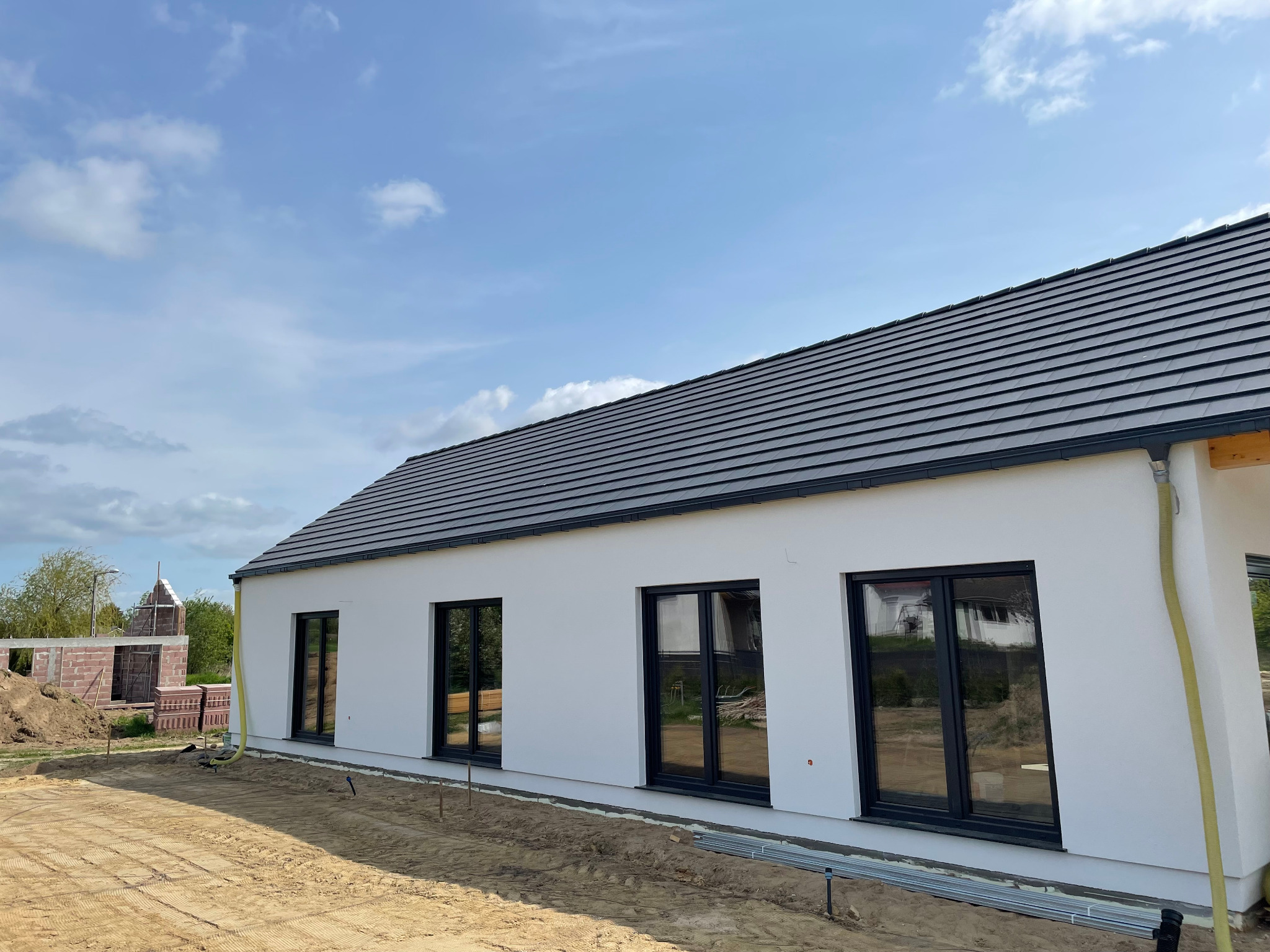 Nowoczesny dom z białymi ścianami i ciemną dachówką, widoczny z zewnątrz na tle błękitnego nieba. W tle widać elementy konstrukcyjne innego budynku.