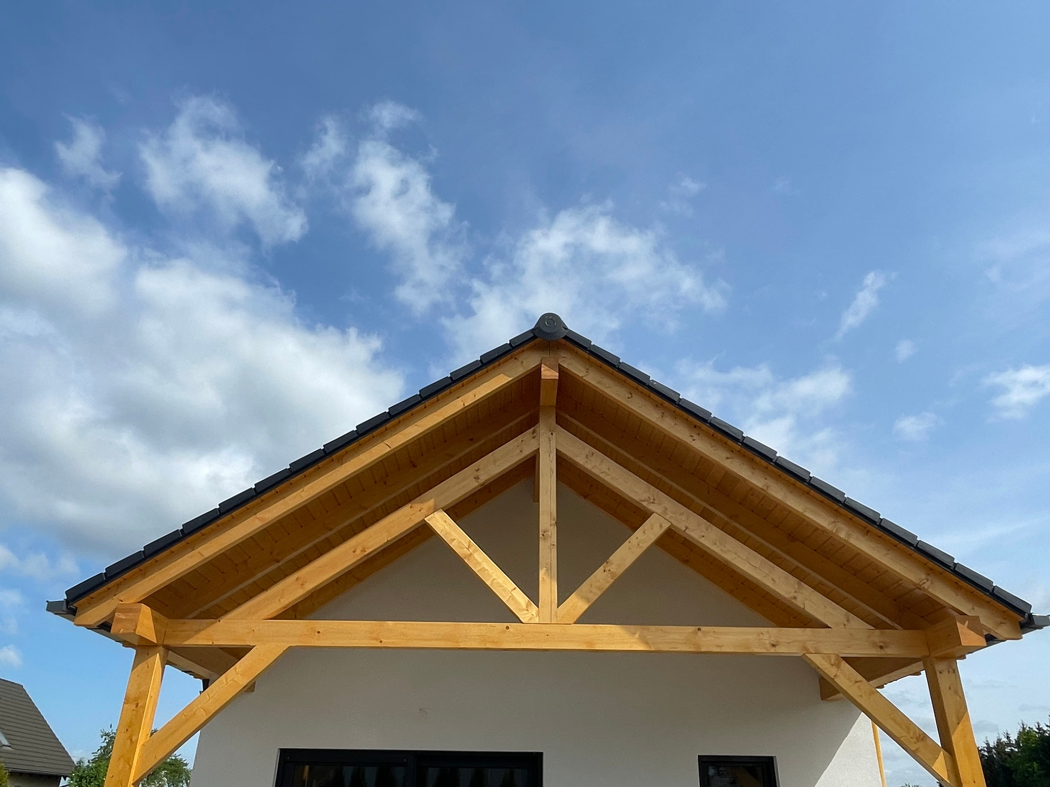 Drewniana konstrukcja dachu z czarną dachówką na tle błękitnego nieba z chmurami. Widoczny fragment białej ściany budynku z oknami.