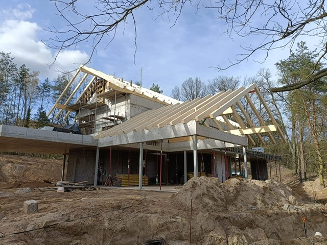 Surowy stan domu z betonu i drewna w trakcie budowy. Widoczna konstrukcja dachu z drewnianych belek na tle drzew i błękitnego nieba.