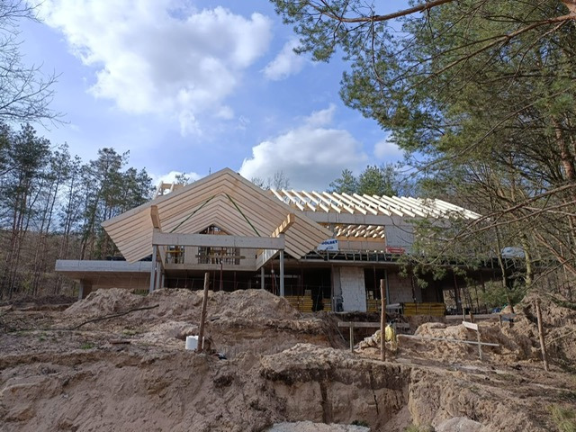 Wznoszenie konstrukcji domu z drewnianą więźbą dachową w Deszcznie. Widok na szkielet budynku w otoczeniu drzew i piaszczystego terenu.