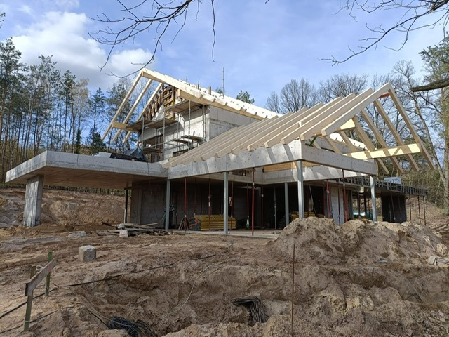 Surowy dom z betonu komórkowego w trakcie budowy, z widoczną drewnianą konstrukcją dachu i podporami. Otoczenie leśne, ziemisty teren budowy.