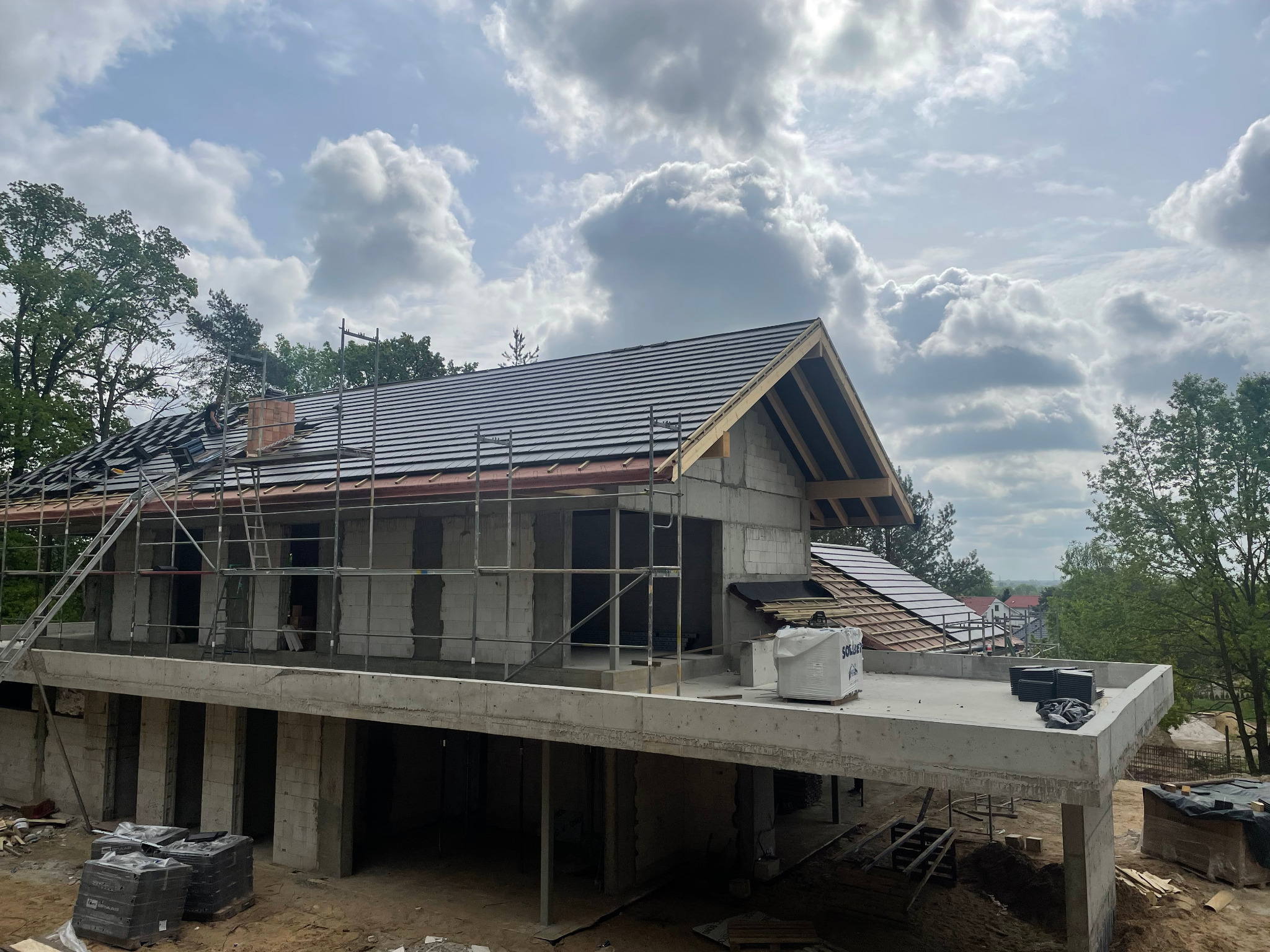 Budowa domu w trakcie: murowana konstrukcja, częściowo pokryty dach z czarnej dachówki, rusztowania. Widok z zewnątrz, w tle zieleń i pochmurne niebo.