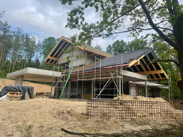 Budowa domu w stanie surowym otwartym: betonowa konstrukcja, częściowo pokryty dach z czarną dachówką, rusztowania, zbrojenia. Otoczenie leśne.