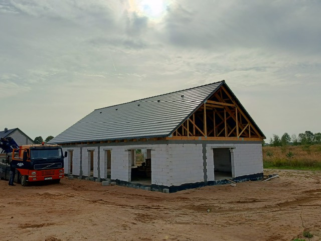 Budowa domu w stanie surowym otwartym: białe ściany z pustaków, drewniana więźba dachowa i szara dachówka. Na pierwszym planie ciężarówka z dźwigiem.