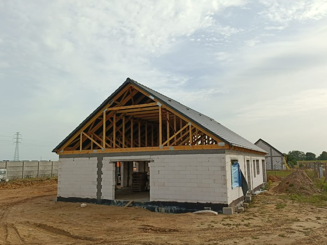 Budowa domu z bloczków: widoczna więźba dachowa z częściowo położonym pokryciem. W tle drugi budynek w trakcie budowy i słup wysokiego napięcia.