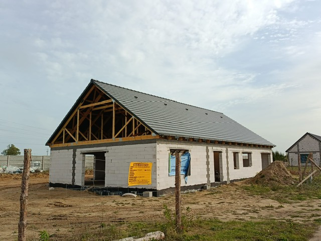 Niedokończony dom z szarej dachówki, widoczna więźba dachowa i ściany z białych bloczków. Na pierwszym planie drewniany słupek ogrodzeniowy i żółta tablica informacyjna.