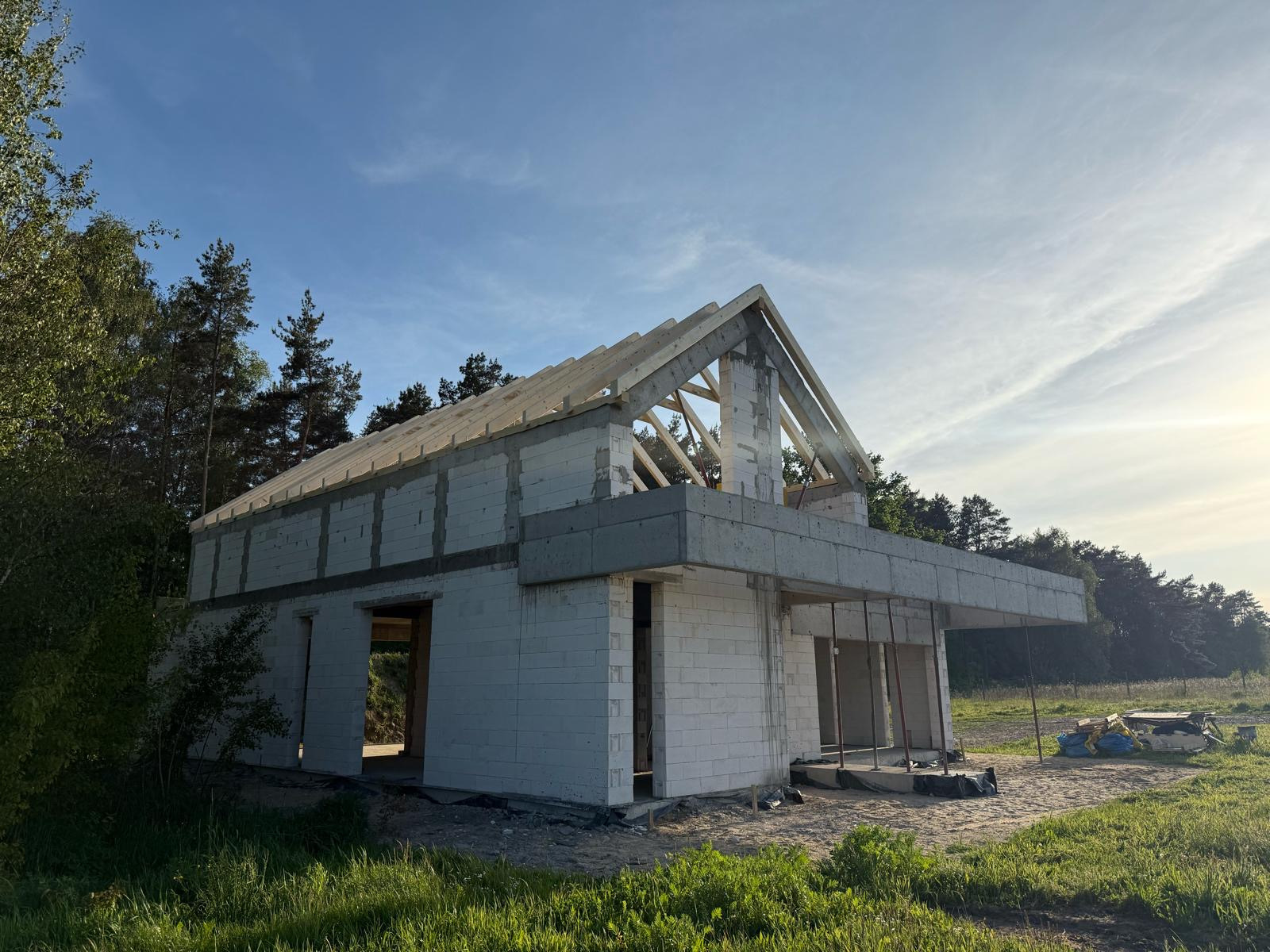 Surowy stan domu z białych bloczków, z widoczną drewnianą konstrukcją dachu i betonowym zadaszeniem nad wejściem, na tle zieleni i błękitnego nieba.