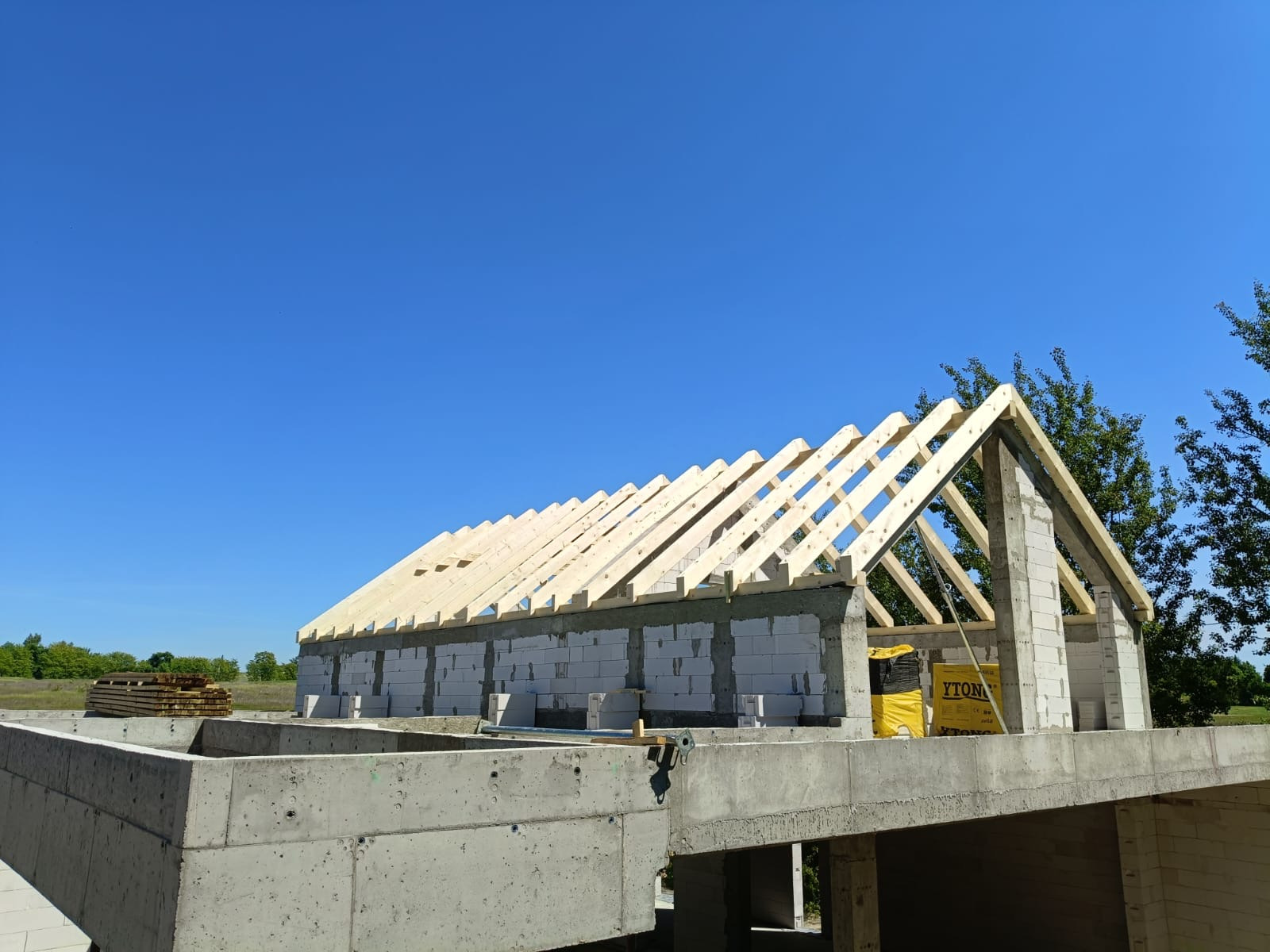 Wznoszenie więźby dachowej na budynku z betonu komórkowego w słoneczny dzień. Widoczne drewniane elementy konstrukcyjne i bloczki Ytong.