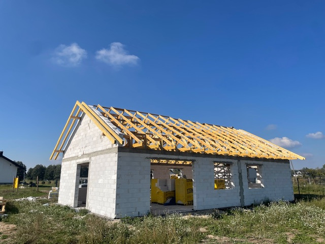 Wznoszenie domu z bloczków: surowe ściany i drewniana więźba dachowa pod błękitnym niebem. Widoczne żółte paczki materiałów budowlanych wewnątrz budynku.