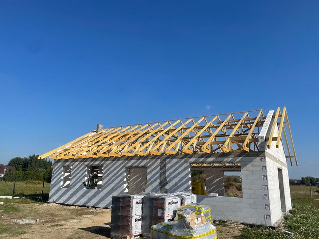 Wznoszenie domu z pustaków: surowy stan z drewnianą konstrukcją dachu i paletami dachówek Roben na pierwszym planie w słoneczny dzień.