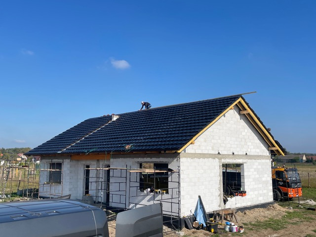 Budowa domu z białego bloczka: czarny dach z dachówki, widoczna więźba, pracownik na dachu. W tle błękitne niebo i zabudowa. Na pierwszym planie fragment auta.