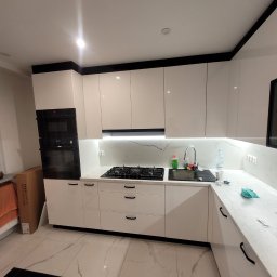 Biała, nowoczesna kuchnia z czarnymi akcentami: fronty na wysoki połysk, marmurowy blat, płyta gazowa i piekarnik w zabudowie. Minimalistyczny design.