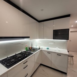 Biała, błyszcząca kuchnia z czarnymi akcentami: okap Amica, płyta gazowa, marmurowy backsplash i podłoga. Nowoczesny design z oświetleniem LED pod szafkami.