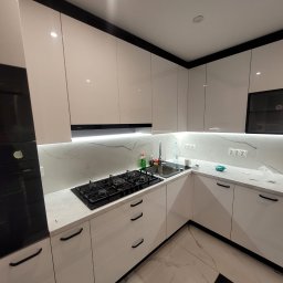 Nowoczesna kuchnia z białymi, lakierowanymi frontami, czarnymi akcentami i marmurowym blatem. Widoczna płyta gazowa i oświetlenie LED pod szafkami.