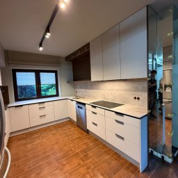 Piotr Warulik - Jasna kuchnia z drewnianymi akcentami i oświetleniem punktowym. Widoczne szafki, blat, okno z żaluzjami i fragment podłogi z płytek imitujących drewno. W lustrze odbija się osoba.