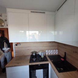 Piotr Warulik - Nowoczesna kuchnia z białymi, błyszczącymi szafkami i drewnianym blatem. Na blacie kuchenka gazowa z garnkami i czarny zlew. Widoczny fragment osoby wchodzącej do pomieszczenia.