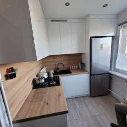 Piotr Warulik - Nowoczesna kuchnia z białymi szafkami i drewnianym blatem. Na blacie kuchenka, garnki i akcesoria. Lodówka w zabudowie. Jasne płytki na podłodze i ścianie.