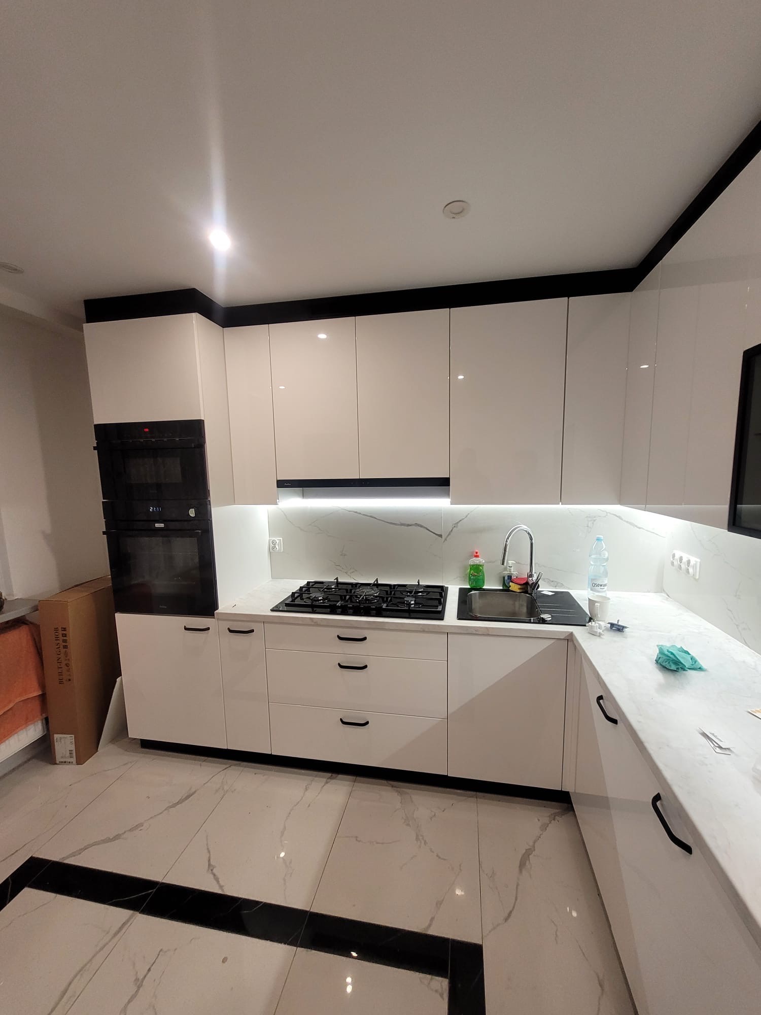 Biała, błyszcząca kuchnia z czarnymi akcentami: fronty szafek, listwy podszafkowe i uchwyty. Marmurowy blat i backsplash. Płyta gazowa i piekarnik w zabudowie.