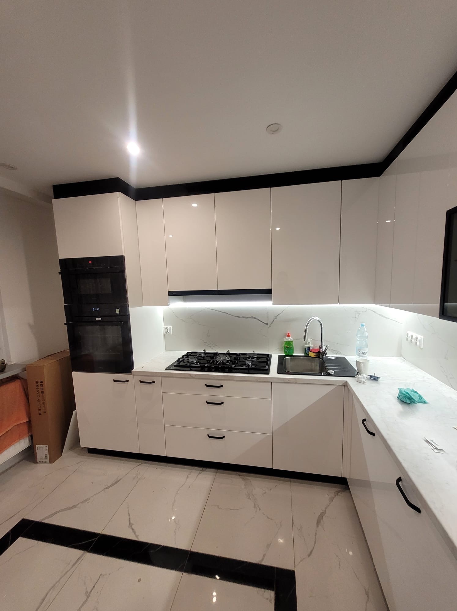 Biała, nowoczesna kuchnia z czarnymi akcentami: fronty na wysoki połysk, marmurowy blat, płyta gazowa i piekarnik w zabudowie. Minimalistyczny design.