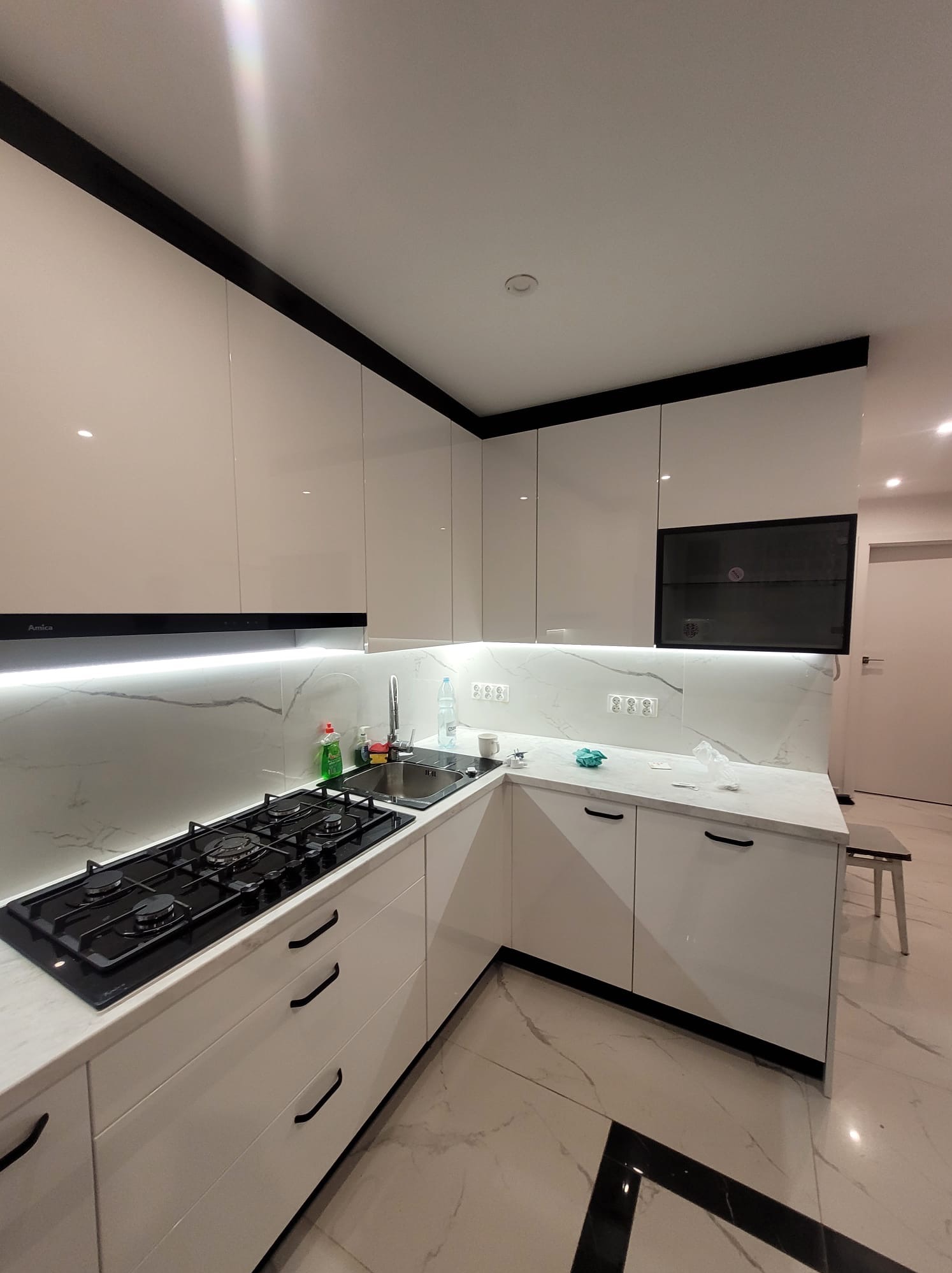 Biała, błyszcząca kuchnia z czarnymi akcentami: okap Amica, płyta gazowa, marmurowy backsplash i podłoga. Nowoczesny design z oświetleniem LED pod szafkami.
