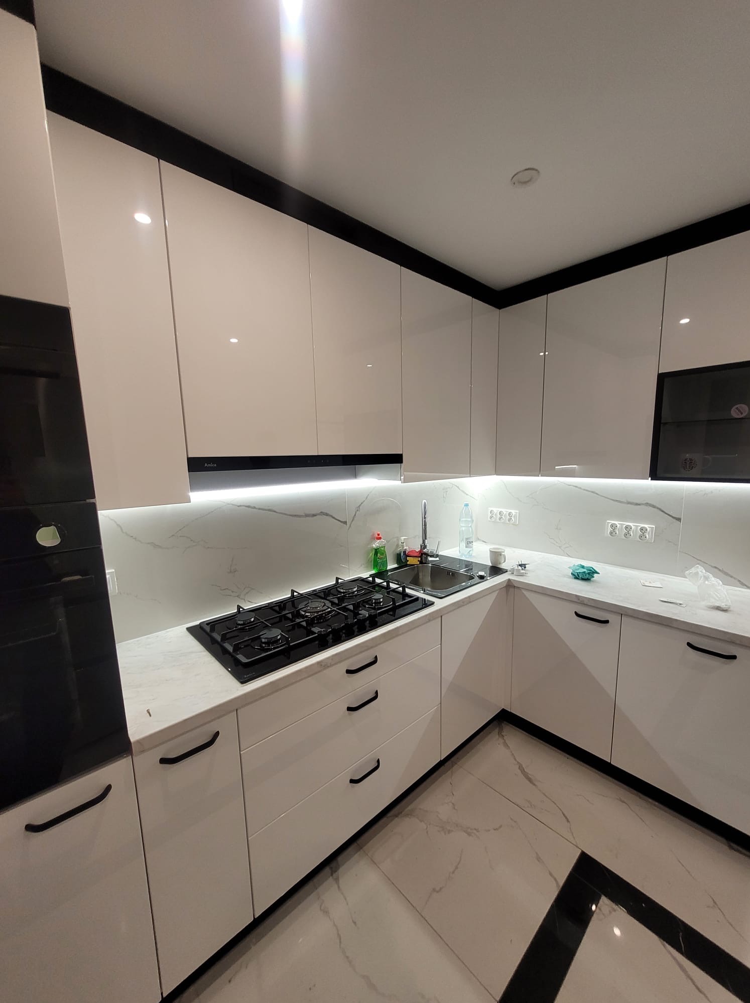 Nowoczesna kuchnia z białymi, lakierowanymi frontami, czarnymi akcentami i marmurowym blatem. Widoczna płyta gazowa i oświetlenie LED pod szafkami.