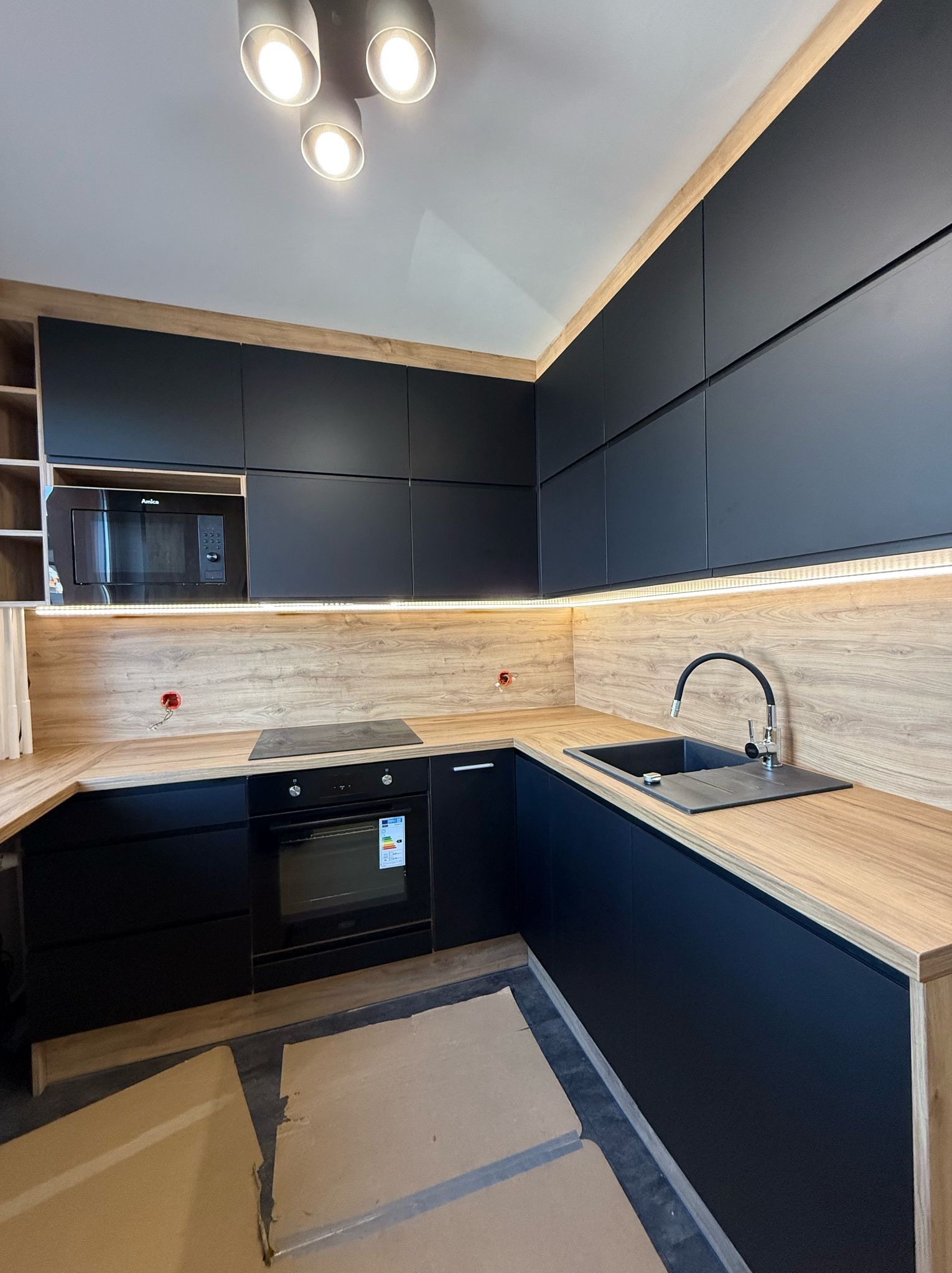 Nowoczesna kuchnia na wymiar w Tychach. Czarne fronty szafek, drewniany blat i podświetlenie LED tworzą elegancki kontrast. Widoczny zlew i płyta indukcyjna.