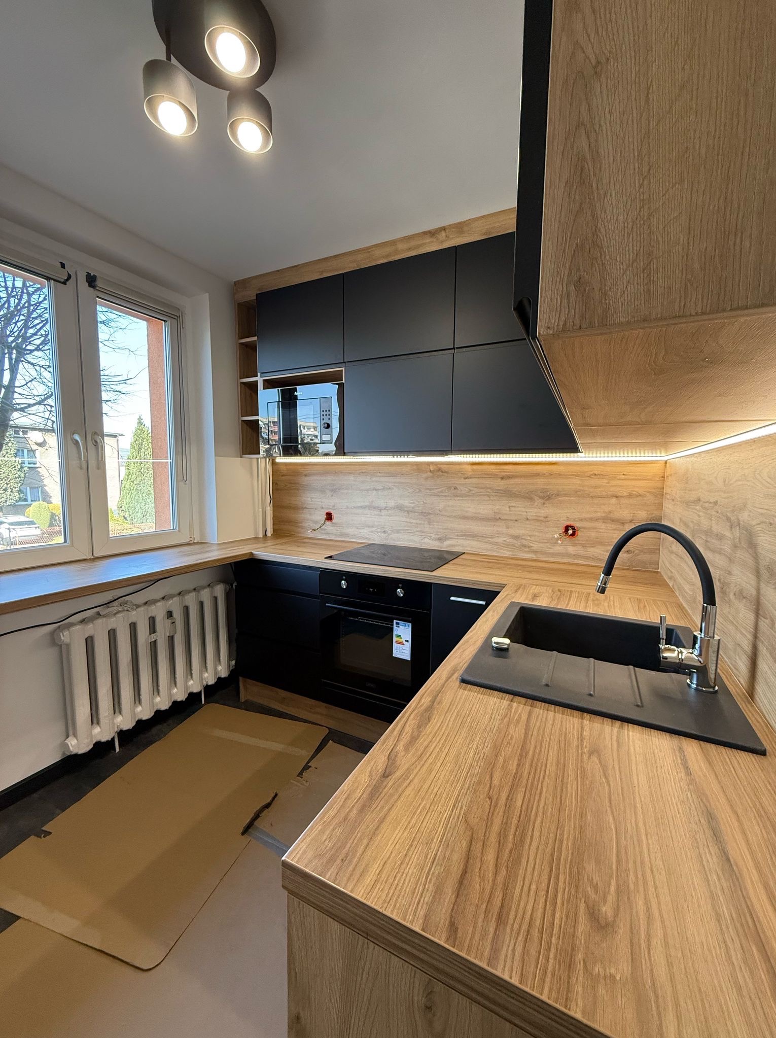 Nowoczesna kuchnia: czarne szafki wiszące, drewniany blat z czarnym zlewem i bateria. Oświetlenie LED pod szafkami. Widok na okno i grzejnik.