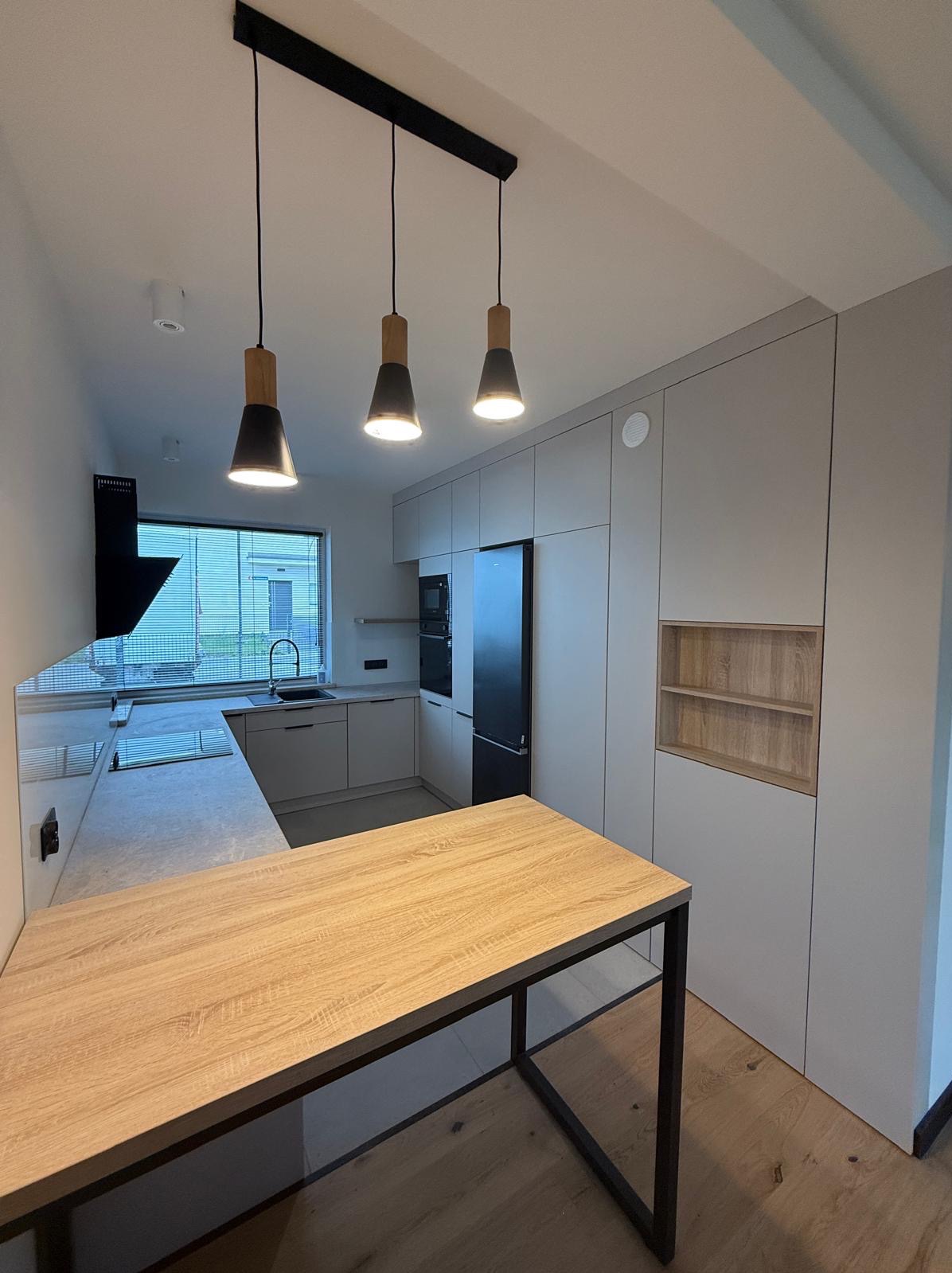 Nowoczesna kuchnia z drewnianym blatem i szarymi szafkami. Nad blatem wiszą trzy lampy z drewnianymi elementami. Widoczne okno i sprzęt AGD.