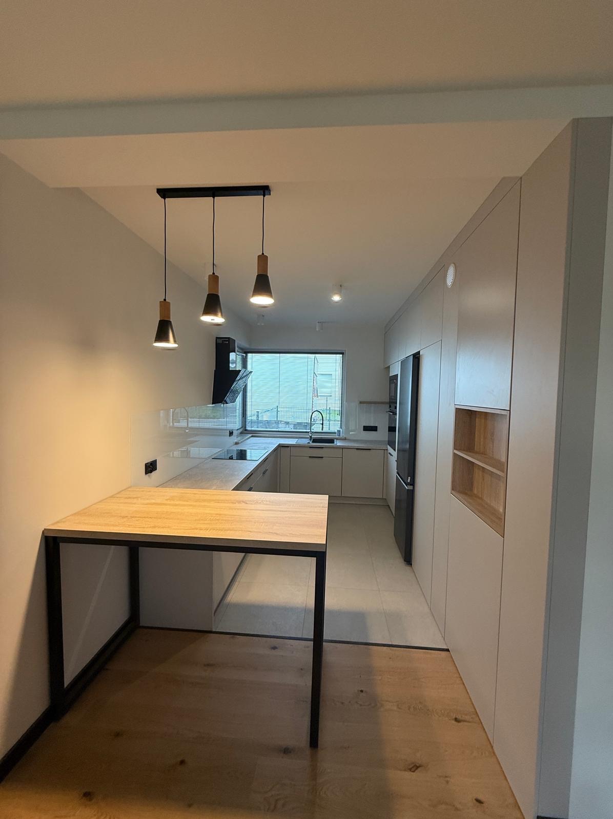 Jasna kuchnia z drewnianym blatem stołu na czarnych nogach. Minimalistyczne lampy wiszące nad blatem. Nowoczesne szafki i sprzęt AGD w tle. Widok na okno.