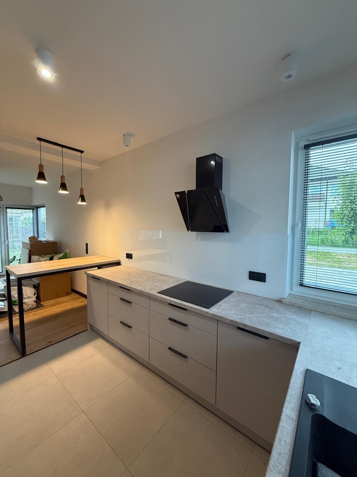 Jasna kuchnia z szarymi frontami i blatem imitującym kamień. Czarny okap nad płytą indukcyjną. Widoczne lampy wiszące i stół z drewnianym blatem na metalowym stelażu.