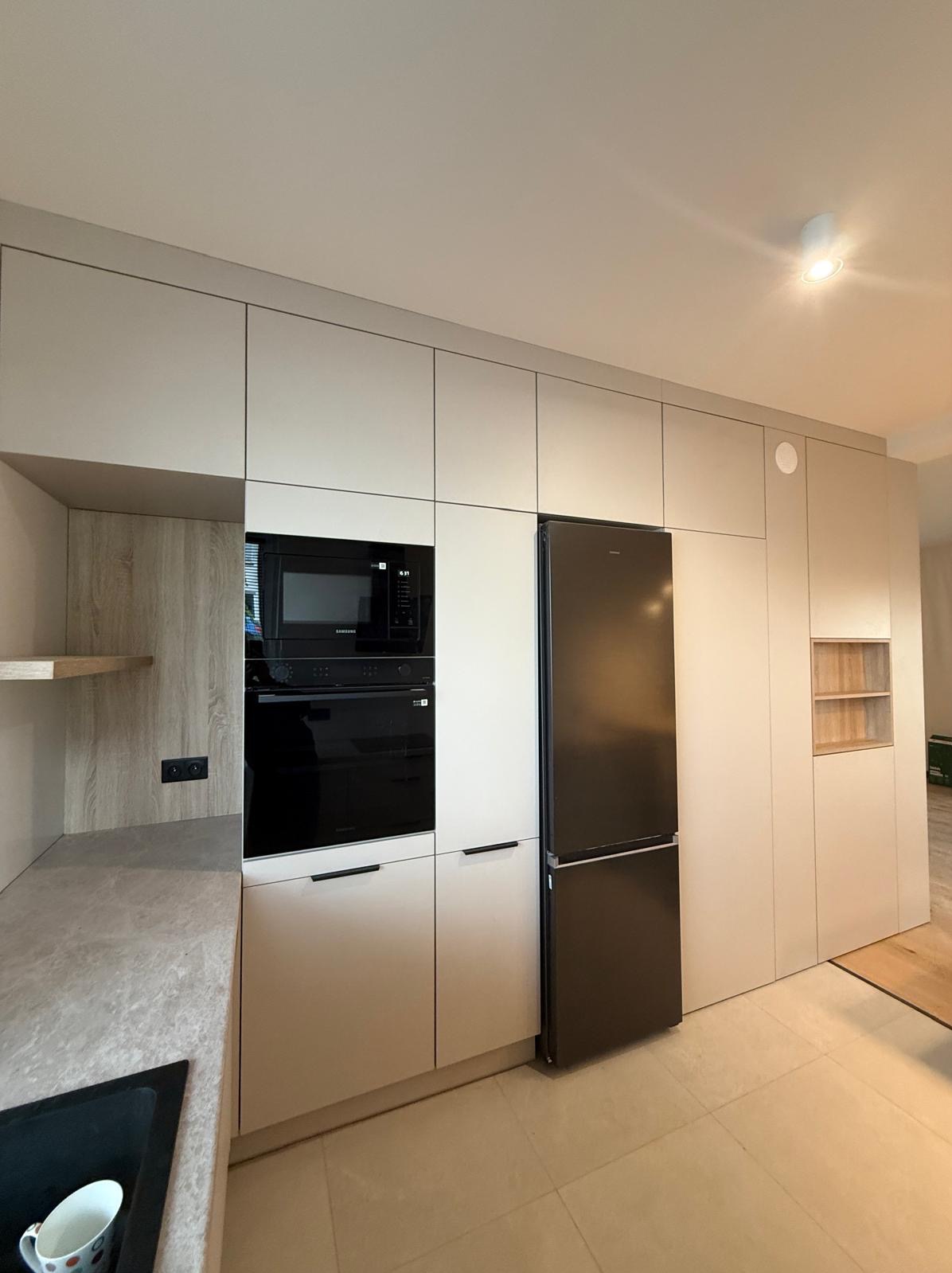 Jasna zabudowa kuchenna z czarnym sprzętem AGD (piekarnik, mikrofalówka, lodówka). Minimalistyczny design, gładkie fronty, długie uchwyty. Widok na fragment blatu i podłogi.